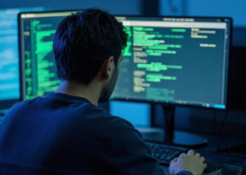 A person with dark hair sits at a desk in front of two computer monitors displaying green code in a dimly lit room, suggesting programming or cybersecurity work such as configuring a personal firewall. | TRWHO.com
