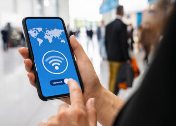 A person holds a smartphone displaying a Wi-Fi connection screen with a world map background and a "Connect" button, while standing in a blurred indoor public space. | TRWHO.com